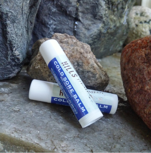 Two tubes of Hills' Cold Sore Balm on a stone surface with rocks in the background